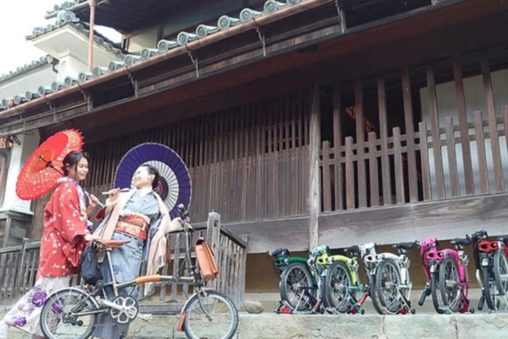 Two women in traditional Japanese clothing holding umbrellas next to parked folding cycles in front of a historic building on a Wakimachi Udatsu bike tour.