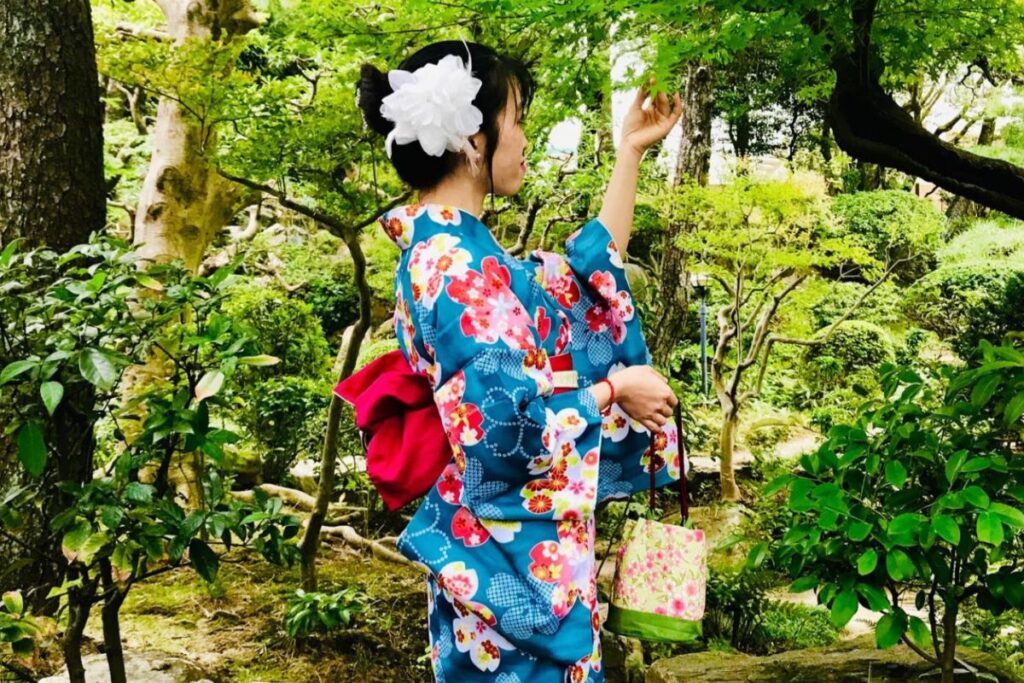 Side profile of a woman in a blue floral-patterned kimono standing in a dense, lush traditional Japanese garden with green foliage, reaching up to touch a leaf.
