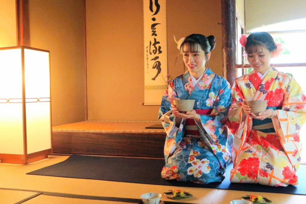 Two women participating in the Kimono and matcha experience Nagoya inside a historic tatami room, holding tea bowls at the Tamezaburo Memorial Museum.