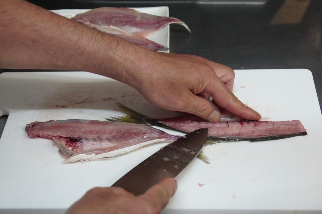 Close-up of a chef demonstrating how to slice a fresh whole aji fish using a professional knife during a Nagoya sushi making masterclass.